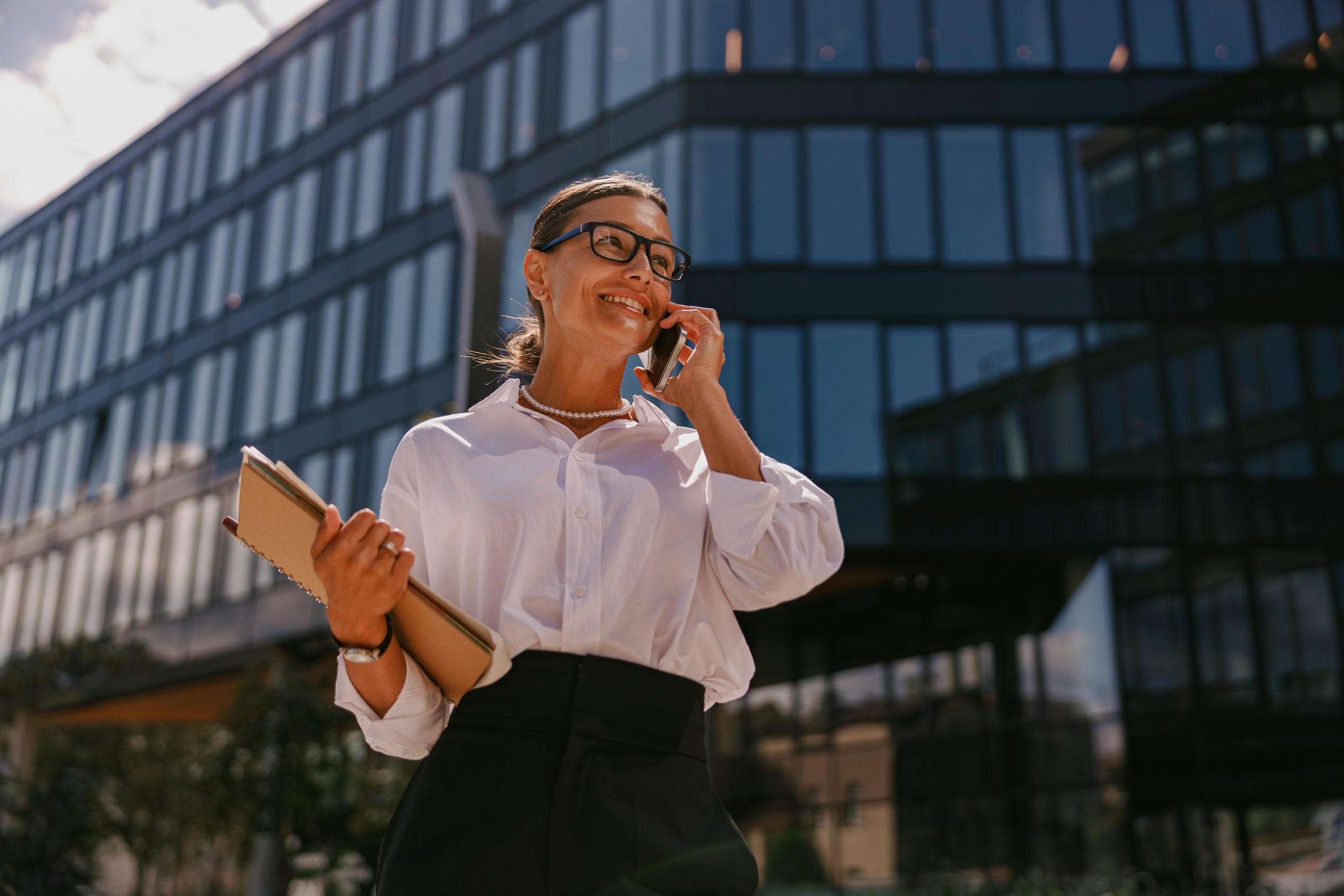 A confident businesswoman on a phone call with a tablet stands in front of a modern office building